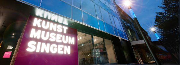 Außenansicht des Kunstmuseum Singen in der Dämmerung; leuchtender Schriftzug, Glasfassade.