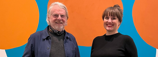 Eine männliche und eine weibliche Person vor einem bunten Kunstwerk blicken auf dne Betrachter und lächeln