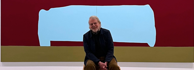 Markus Weggenmann sitzt vor einer seiner großformatigen, farbintensiven Arbeiten und lächet den Betrachter an