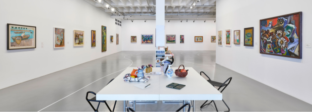 9.	Ausstellungsraum mit zentralem Arbeitstisch und farbigen Gemälden an den umliegenden Wänden. 