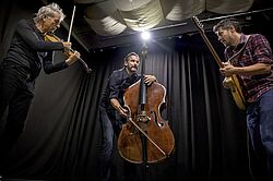 Drei Musiker spielen Violine, Kontrabass und Gitarre auf einer Bühne vor schwarzem Vorhang.