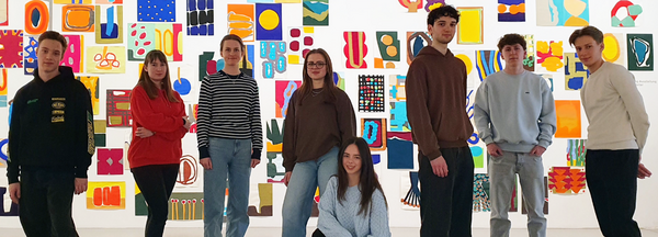 Jugendliche stehen locker in einer Reihe mit dem Blick auf die Betrachtenden gerichtet, im Hintergrund: eine Wand voller bunter Arbeiten auf Papier 